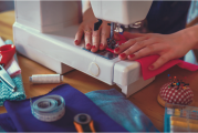 A close up of a woman using a sewing machine. Various sewing items like thread and pin cushions lay in the foreground