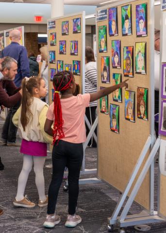 Two look at art display as one points to a portrait.