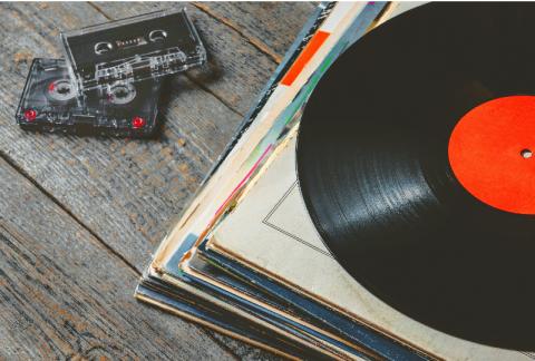 A stack of 2 cassettes on the left side with a stack of several vinyl records on the right. One record sits atop the pile without its cover, the rest of stored