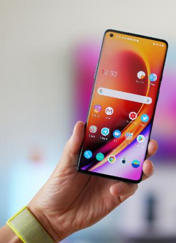 A left hand holding an android smartphone. The phone screen is on, featuring the home screen which has several apps and a google search bar