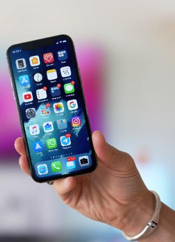 A woman's right hand holding an iPhone. The phone screen is on, featuring the home screen which has dozens of apps on it